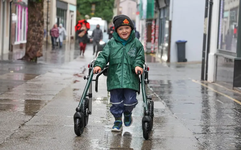 Gregor Corse walking with his walker along a wet street, wearing a green raincoat.