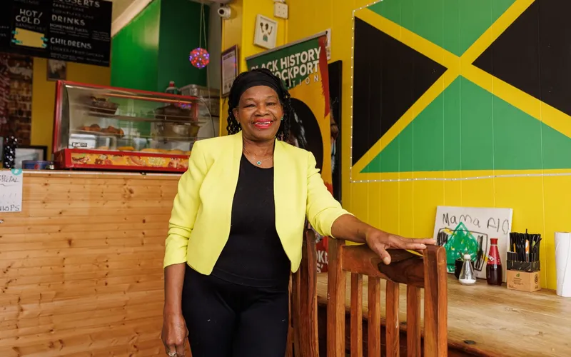 Florence Coke in a yellow jacket inside her takeaway, with a Jamaican flag painted on the wall.